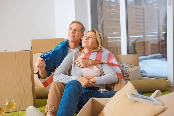 Happy couple wrapped in usa national flag looking up while sitting near cardboard boxes — Stock Photo