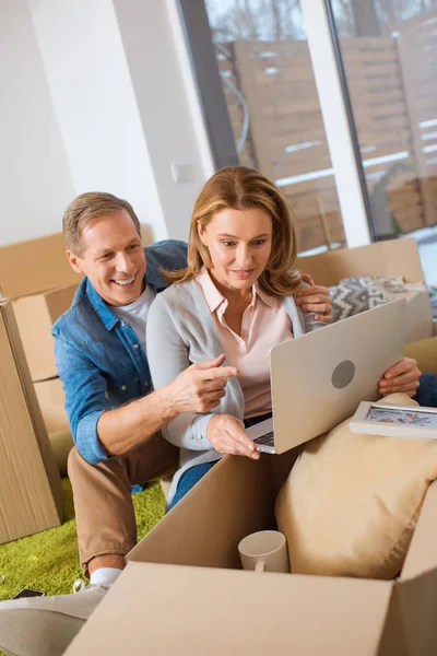 Couple souriant à l'aide d'un ordinateur portable tout en étant assis près des boîtes en carton à la nouvelle maison — Photo de stock