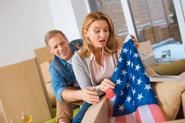 Mujer sorprendida consiguiendo bandera de los E.E.U.U. fuera de la caja de cartón mientras que se sienta cerca de marido sonriente en nuevo hogar - foto de stock