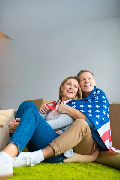 Felice coppia avvolto in USA bandiera nazionale guardando in alto mentre seduto sul tappeto verde a nuova casa — Foto stock