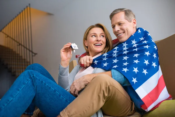 Mujer feliz sosteniendo llaves con baratija modelo de casa mientras envuelve en bandera nacional de EE.UU. con el marido - foto de stock