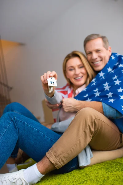 Fuoco selettivo della donna felice che tiene le chiavi con il gingillo del modello della casa mentre avvolge in bandiera nazionale degli Stati Uniti con il marito — Foto stock