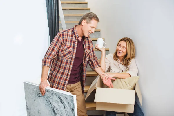 Усміхнений чоловік тримає фотографію, стоячи біля дружини, сидячи на сходах і розпаковуючи картонну коробку — стокове фото