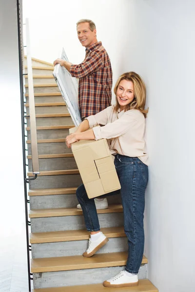 Sonriente pareja llevando cosas arriba y mirando a la cámara - foto de stock