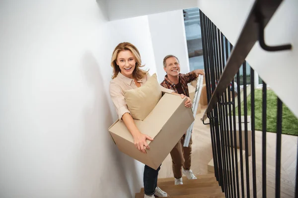 Sonriendo feliz pareja llevando cosas arriba y mirando a la cámara - foto de stock