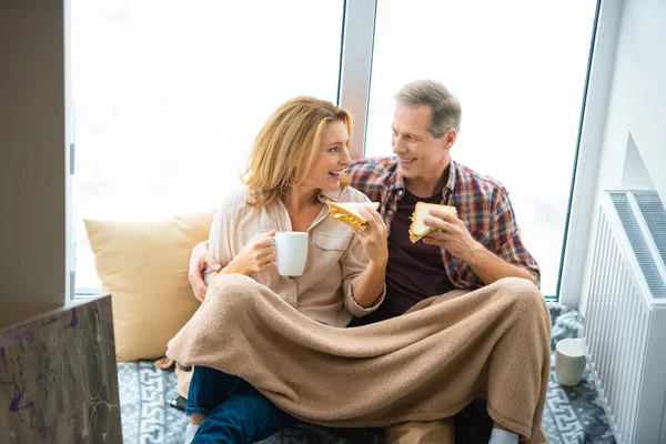 Happy couple eating sandwiches and drinking coffee while sitting by window at new home — Stock Photo