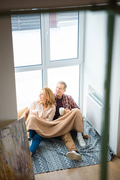 Happy couple sitting on floor by large window and holding coffee cups — Stock Photo