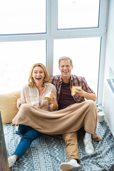 Laughing couple eating sandwiches and drinking coffee while on floor by large window — Stock Photo