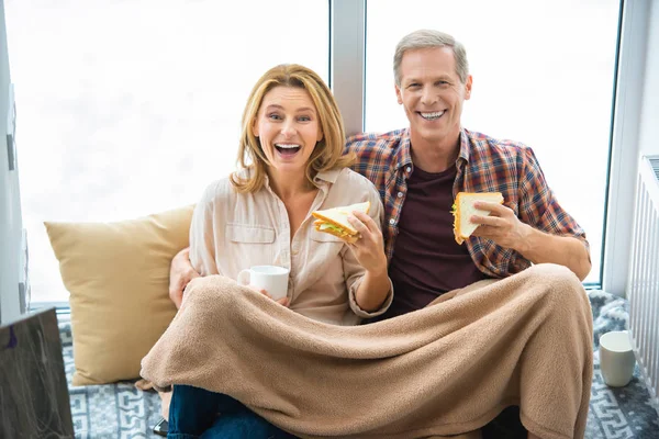 Happy couple eating sandwiches while sitting on floor by large window — Stock Photo