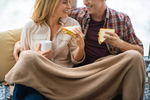 Happy couple eating sandwiches and looking at each other while sitting under soft fleece blanket — Stock Photo