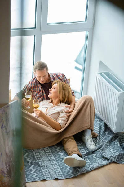 Feliz pareja con copas de vino blanco descansando en el suelo por una gran ventana en el nuevo hogar - foto de stock