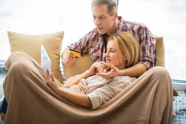 Frau benutzt Laptop mit Ehemann, der Kreditkarte hält, während sie unter weicher Fleece-Decke ruht — Stockfoto