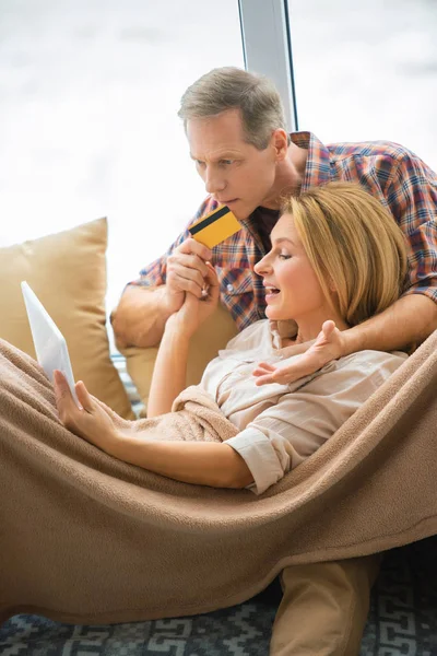 Femme utilisant un ordinateur portable tout mari tenant la carte de crédit à la maison — Photo de stock