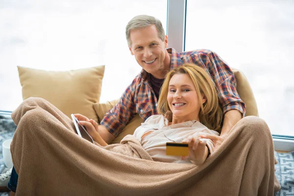 Selective focus of woman showing credit card and holding digital tablet while resting with husband under soft fleece blanket — Stock Photo