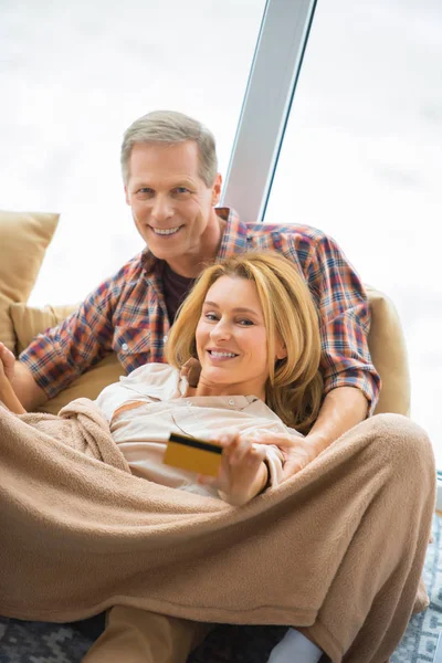 Selektiver Fokus der hübschen Frau, die ihre Kreditkarte zeigt, während sie sich mit ihrem Mann durch ein großes Fenster ausruht — Stockfoto