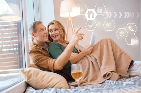 Feliz esposa sosteniendo tableta digital mientras descansa con el marido bajo manta de lana acogedor, concepto de hogar inteligente - foto de stock