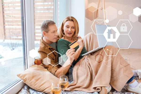 Щасливий чоловік тримає кредитну картку, використовуючи ноутбук разом з дружиною, концепція розумного будинку — стокове фото