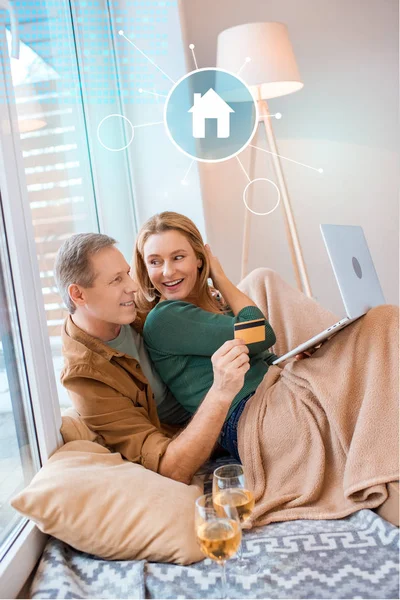 Hombre que sostiene la tarjeta de crédito mientras que usa el ordenador portátil junto con la esposa sonriente, concepto de hogar inteligente - foto de stock