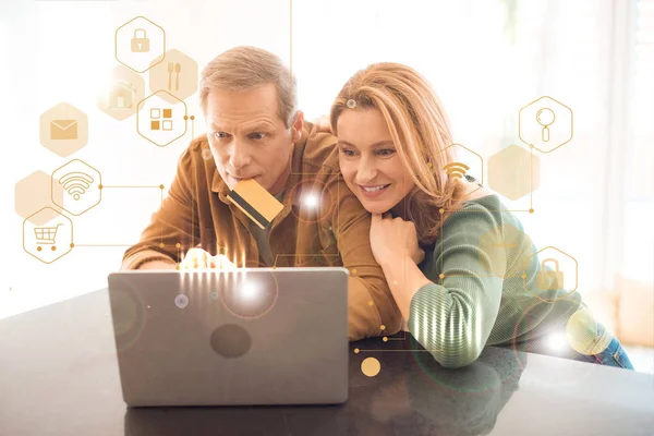 Concentrated man holding credit card with lips while using laptop together with smiling wife, smart home concept — Stock Photo