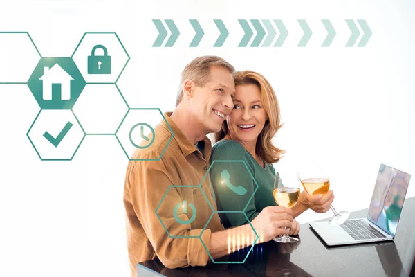 Happy couple holding glasses of white wine while standing by table with laptop, smart home concept — Stock Photo