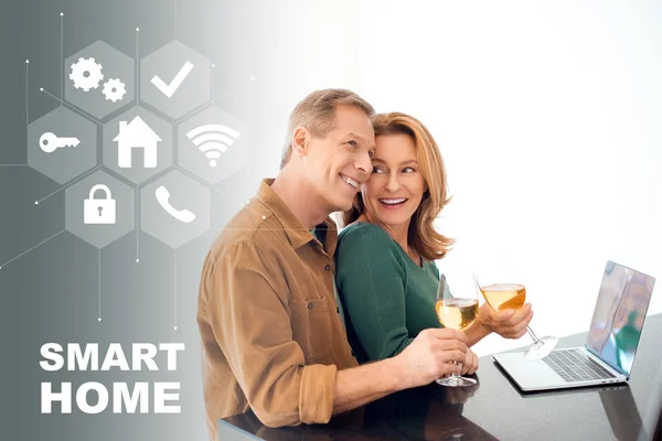 Feliz pareja sosteniendo vasos de vino blanco mientras está de pie a la mesa con el ordenador portátil, concepto de hogar inteligente - foto de stock