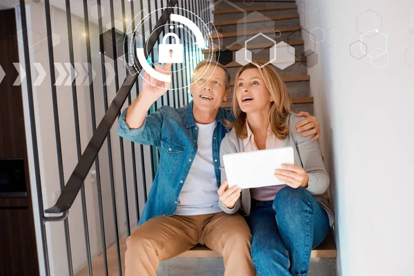 Sonriente hombre apuntando mano y esposa sosteniendo tableta digital mientras está sentado en las escaleras, concepto de hogar inteligente - foto de stock