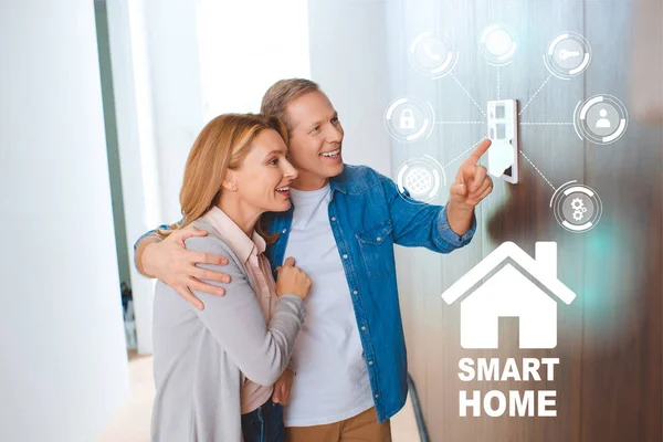 Glücklicher Ehemann umarmt Ehefrau, während er auf Smart House System Control Panel zeigt — Stockfoto
