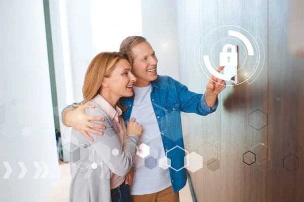 Smiling husband pointing at smart house system control panel and hugging happy wife — Stock Photo