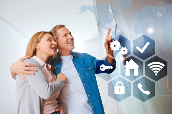 Smiling husband hugging happy wife while pointing at smart house system control panel — Stock Photo