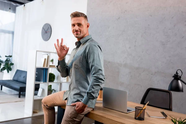 Щасливий бізнесмен, показуючи знак ОК, сидячи з рукою в кишені в офісі — стокове фото
