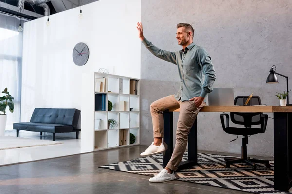 Веселий бізнесмен махає рукою і стоїть з рукою в кишені в сучасному офісі — стокове фото