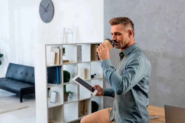 Handsome businessman drinking coffee drom disposable cup and holding digital tablet in office — Stock Photo