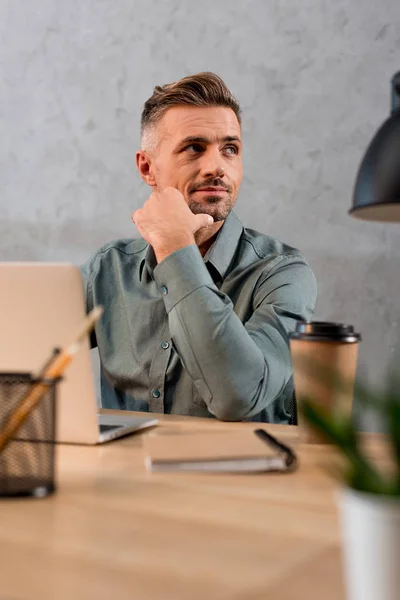 Foco seletivo de homem de negócios pensativo sentado perto do computador portátil no escritório moderno — Fotografia de Stock