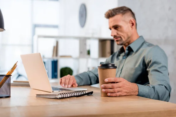 Messa a fuoco selettiva della tazza di carta in mano dell'uomo d'affari che utilizza il computer portatile in ufficio — Foto stock