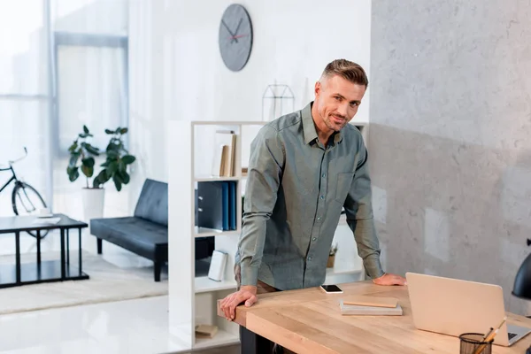 Fröhlicher Geschäftsmann steht in der Nähe seines Arbeitsplatzes im modernen Büro — Stockfoto