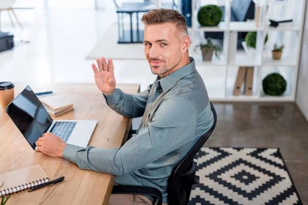 Hombre de negocios feliz utilizando el ordenador portátil con pantalla en blanco mientras mira a la cámara y la mano agitada - foto de stock