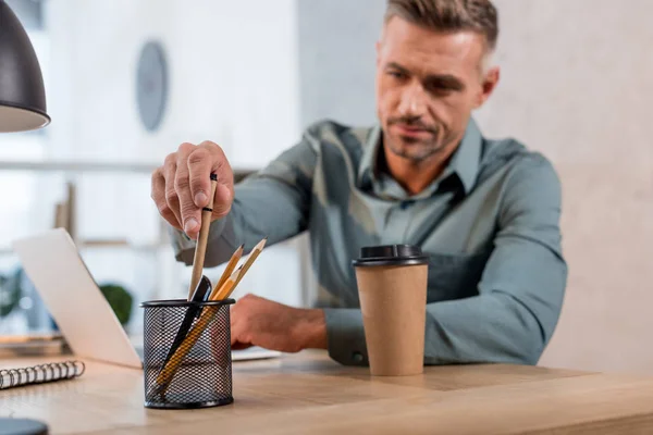 Enfoque selectivo del hombre que toma la pluma del soporte metálico con papelería cerca de la taza de papel en la oficina - foto de stock