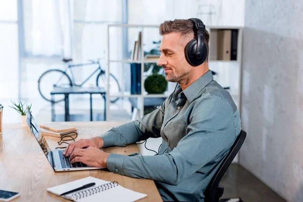 Cheerful businessman listening music in headphones while using laptop in modern office — Stock Photo