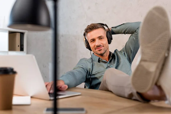 Selektiver Fokus des glücklichen Mannes, der Musik im Kopfhörer hört, während er sich im Büro entspannt — Stockfoto