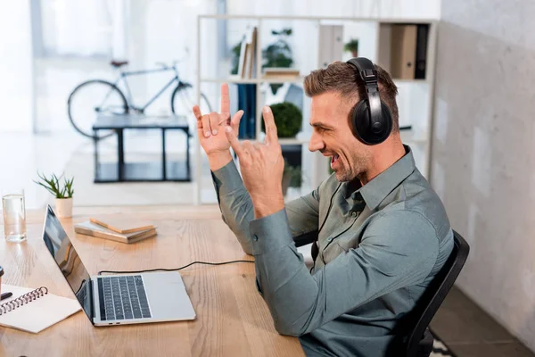 Feliz hombre de negocios escuchando música en los auriculares y mirando a la computadora portátil mientras muestra el cartel de rock en la oficina - foto de stock