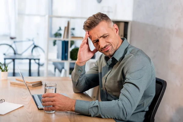 Bel homme tenant un verre d'eau tout en ayant mal à la tête au bureau — Photo de stock