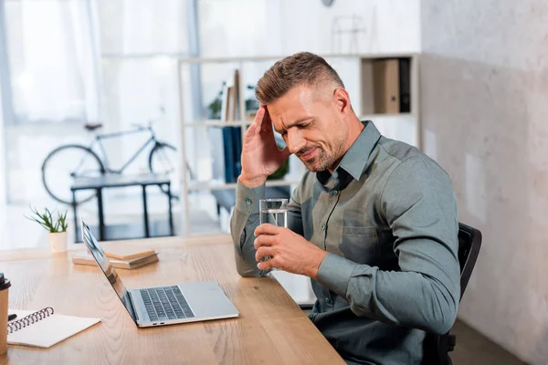 Виснажений бізнесмен тримає склянку води, маючи головний біль в офісі — стокове фото