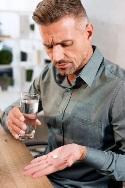 Guapo hombre de negocios mirando píldora mientras sostiene vaso de agua en la oficina - foto de stock