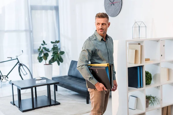 Веселый бизнесмен, стоящий с папками в современном офисе — стоковое фото