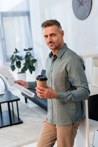 Homme d'affaires joyeux tenant journal et tasse en papier dans les mains tout en regardant la caméra — Photo de stock