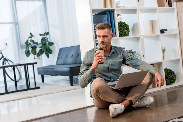 Bonito empresário sentado no chão com laptop e café para ir no escritório — Fotografia de Stock