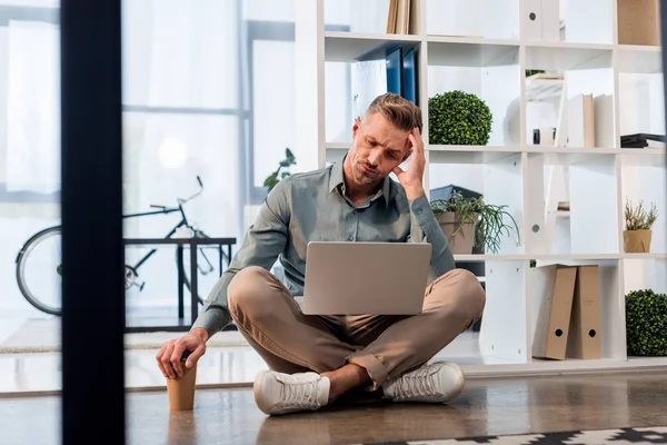 Hombre de negocios guapo sentado con las piernas cruzadas en el suelo y mirando a la computadora portátil - foto de stock