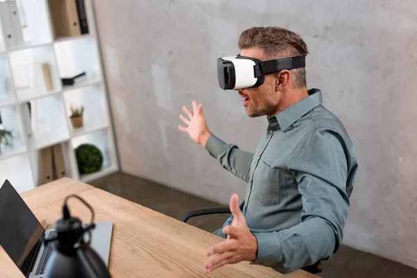 Homem surpreso vestindo fone de ouvido realidade virtual enquanto sentado perto do laptop no escritório — Fotografia de Stock