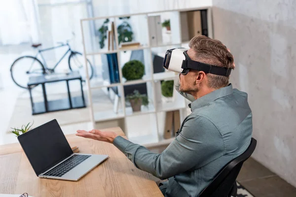 Hombre de negocios con auriculares de realidad virtual mientras está sentado cerca de la computadora portátil con pantalla en blanco - foto de stock