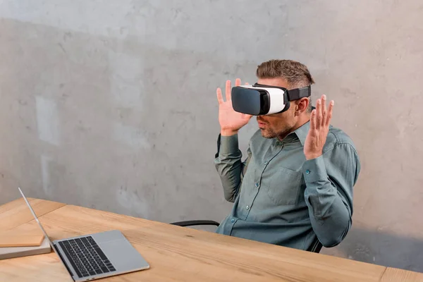 Sorprendido hombre de negocios con auriculares de realidad virtual mientras estaba sentado cerca de la computadora portátil en la oficina - foto de stock